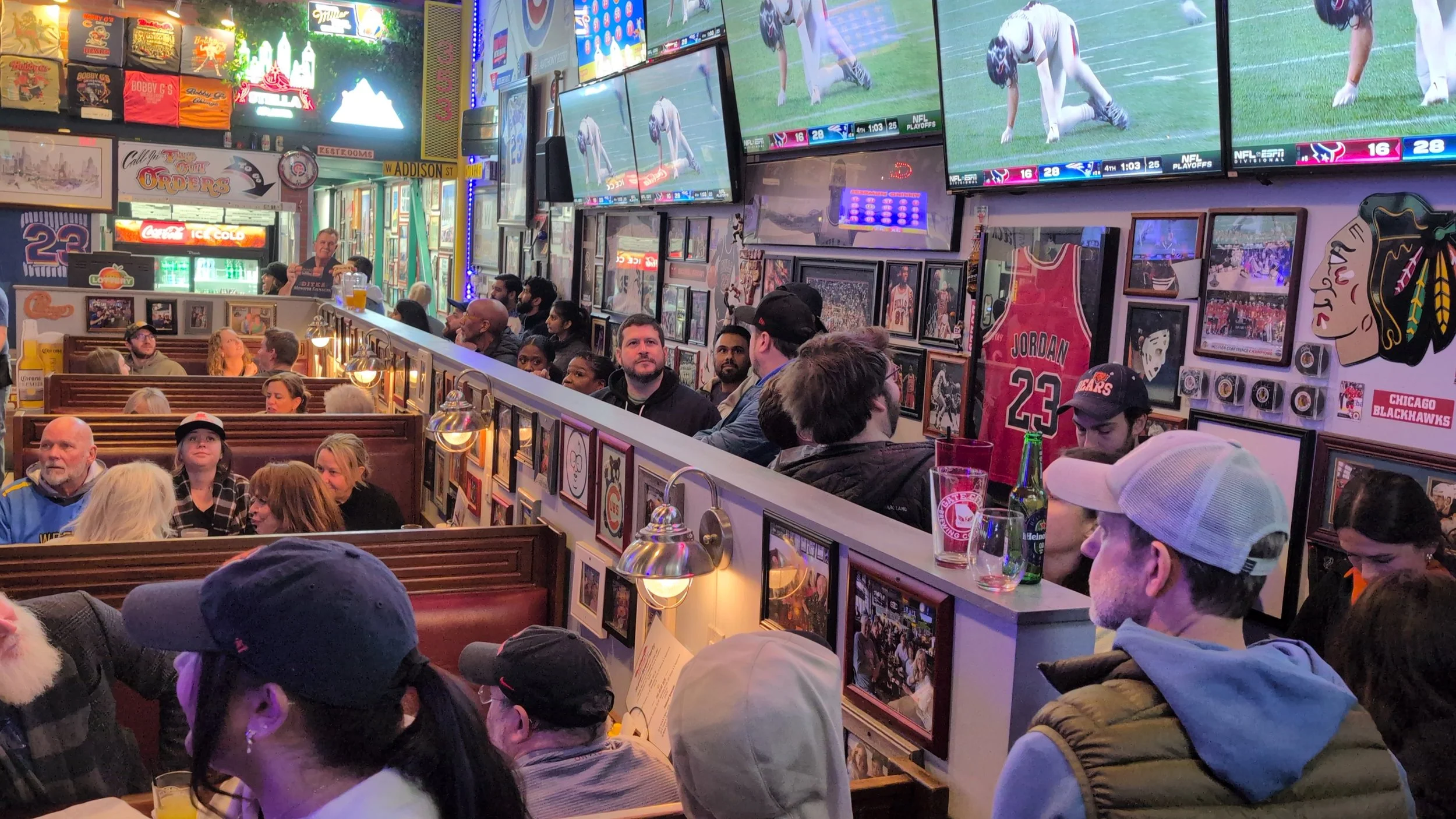 Crowded sports bar filled with people watching multiple flat-screen TVs showing football games, decorated with sports memorabilia, framed photos, jerseys, and colorful signs.
