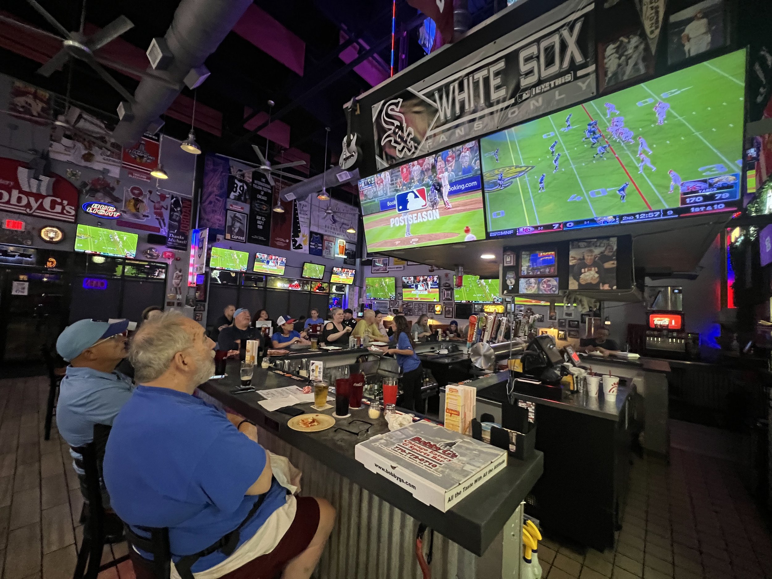 People watching a football game on large TVs at a sports bar. Multiple screens show the game, with some patrons seated at the bar and others at tables.
