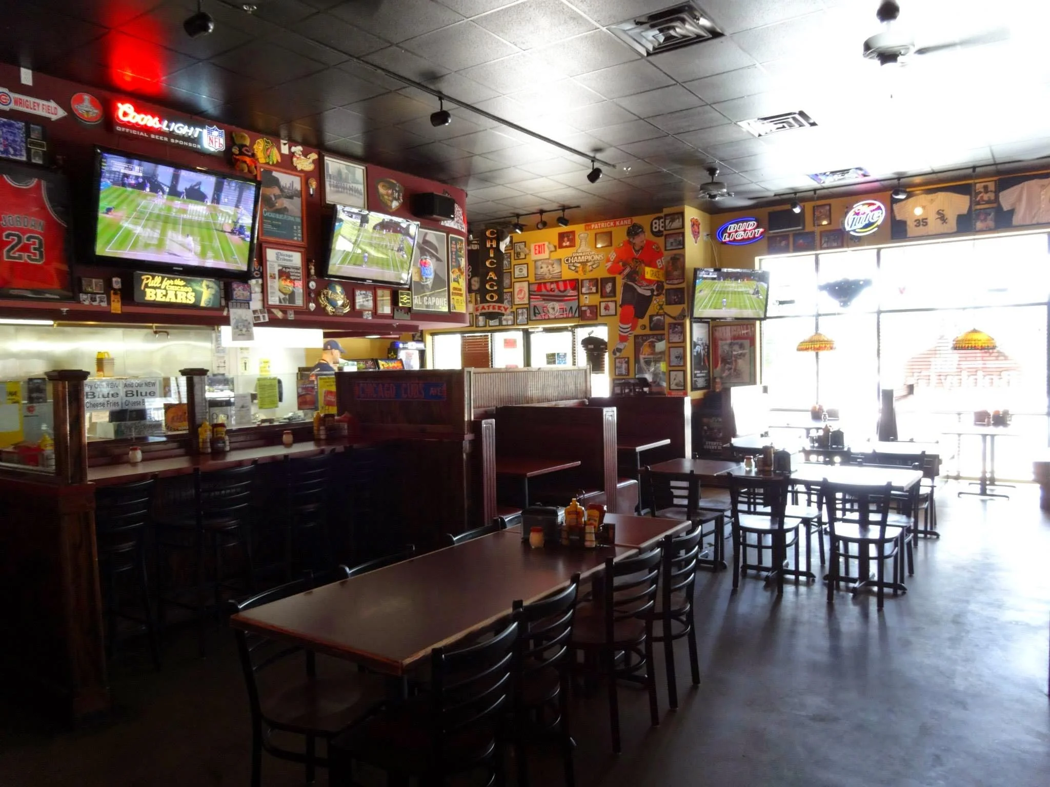 Interior of a sports bar with multiple flat-screen TVs showing a hockey game, decorated with sports memorabilia, jerseys, and framed pictures, with dark wood furniture and tables, and large windows letting in sunlight.