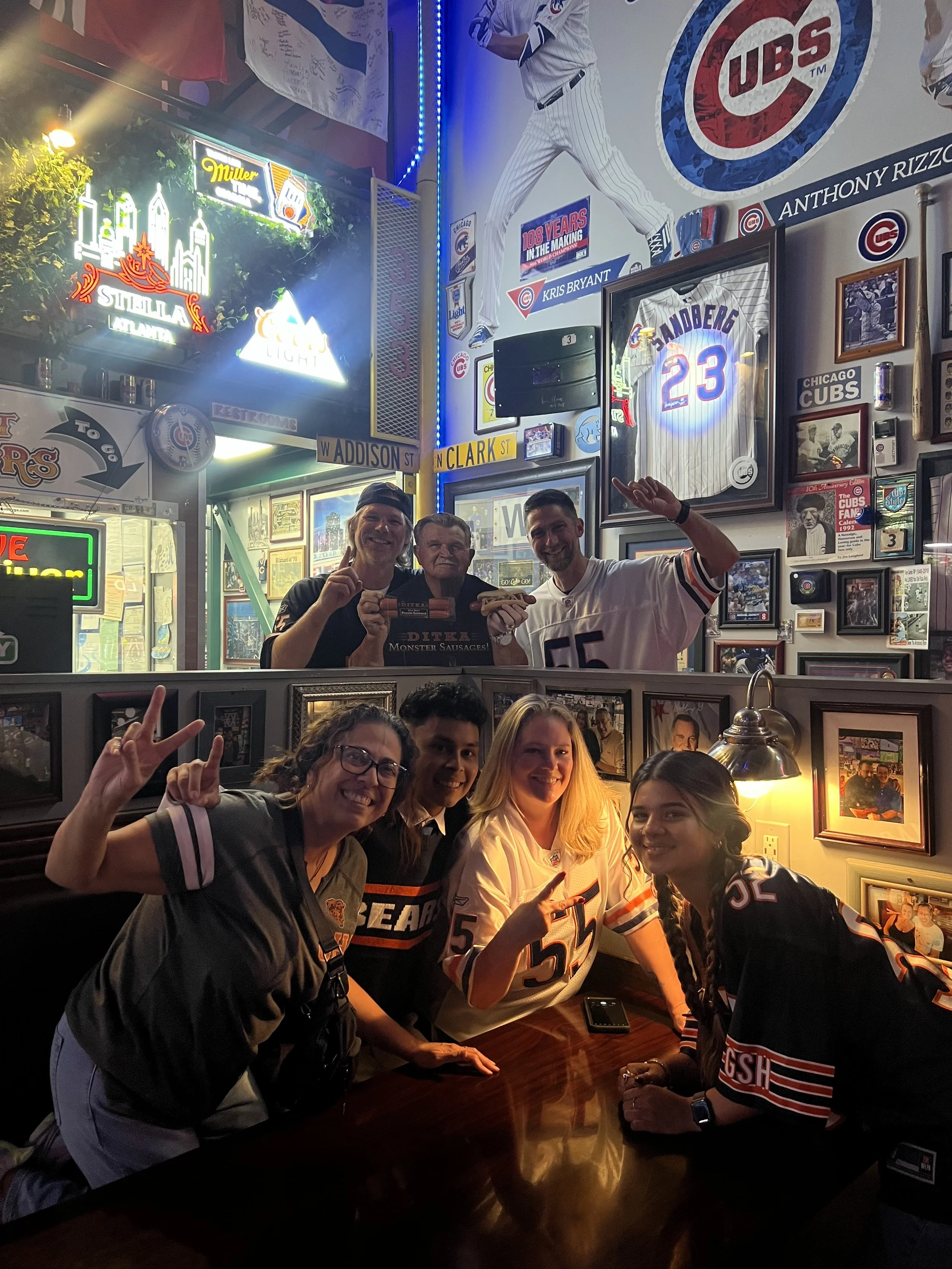 A group of people celebrating at a Chicago Cubs-themed bar, wearing Cubs jerseys, with memorabilia, framed photos, and sports decor on the walls, and a vibrant atmosphere illuminated by neon and warm lighting.
