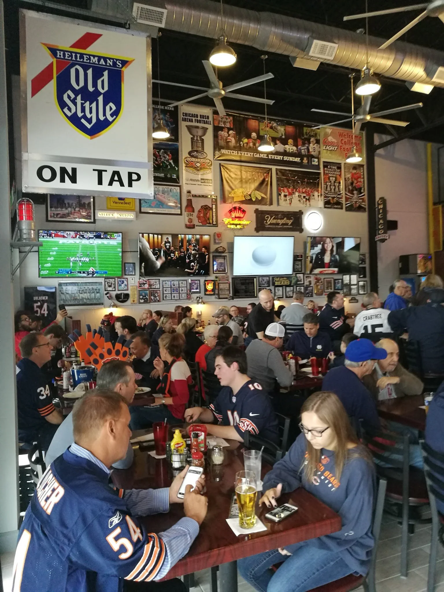 Sports bar filled with people watching a game on multiple televisions, with sports memorabilia, neon signs, and vintage posters on the walls.