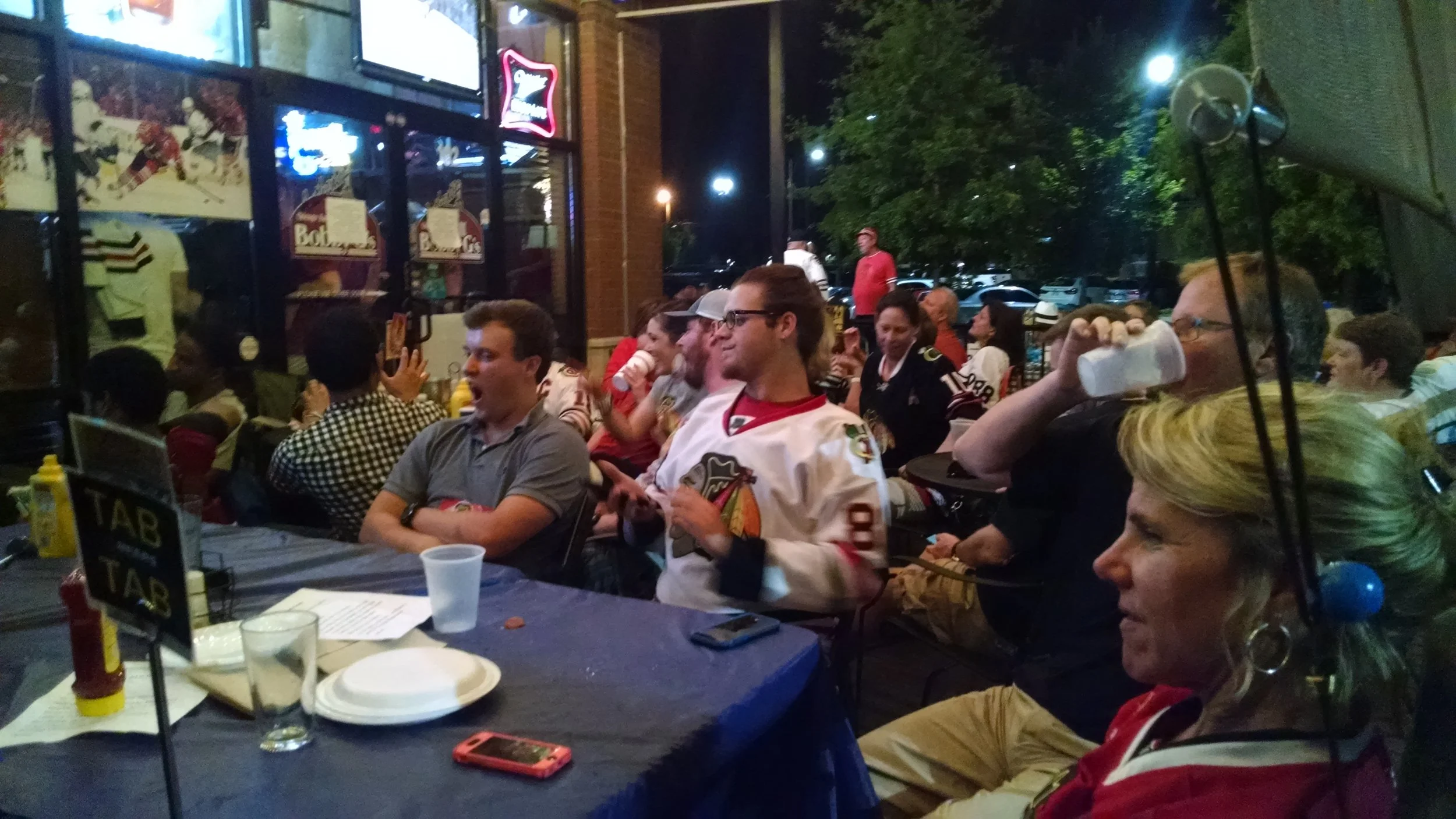 Group of people sitting around a table outside, watching a game on a large screen, some wearing hockey jerseys, with food, drinks, and condiments on the table, at night.
