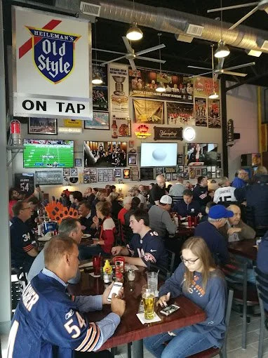 Crowded bar with sports fans, NFL jerseys, people watching games, televisions, neon signs, and a large Hellman's Old Style beer sign.