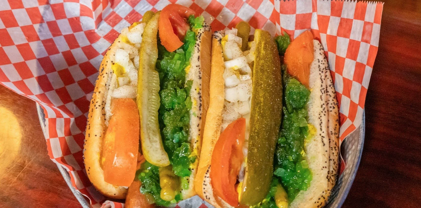 Three hot dogs with chopped onions, pickles, lettuce, tomato, mustard, and relish in a basket lined with orange checkered paper.