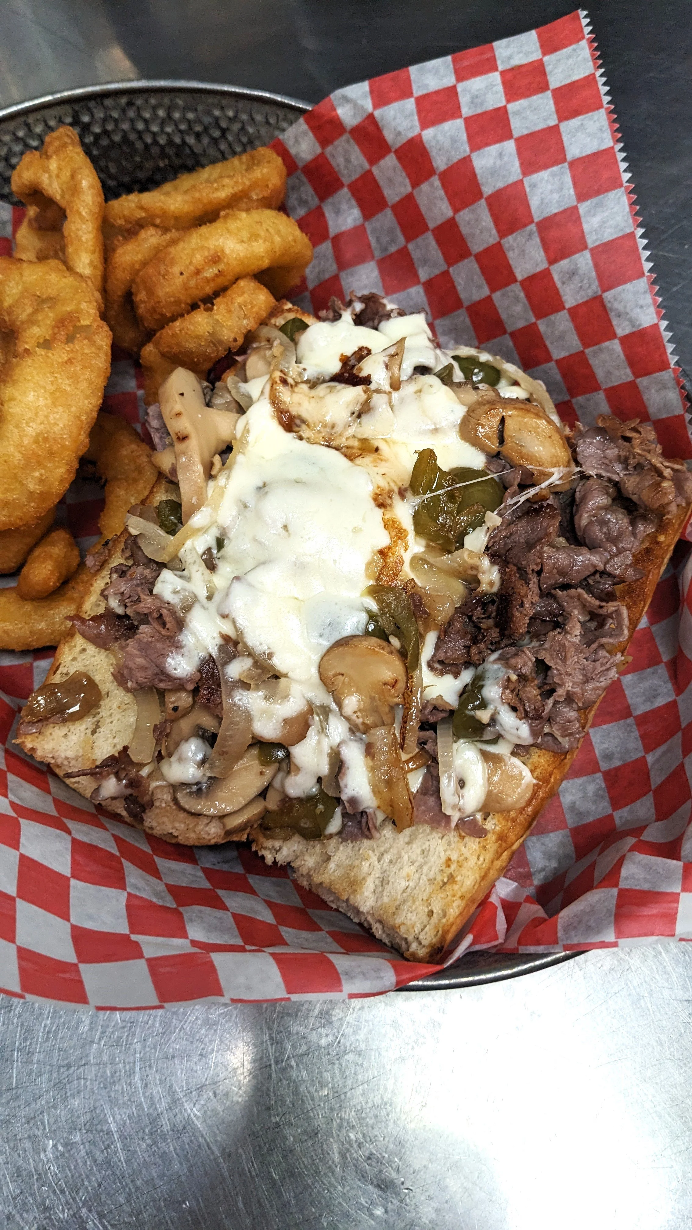 Beef and mushroom hoagie sandwich with French fries on red checkered paper in a metal basket.