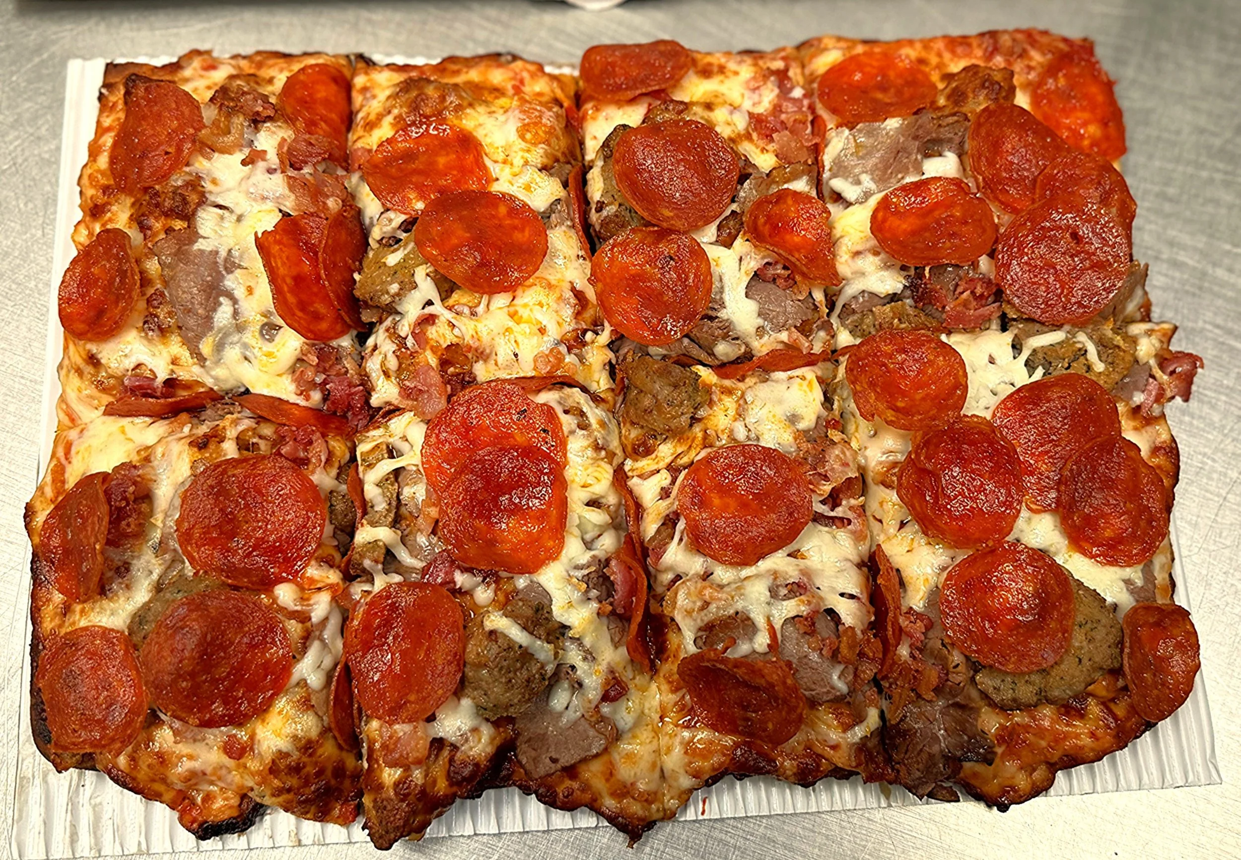 A rectangular pizza with pepperoni slices, sausage, bacon, and melted cheese on a white paper tray.