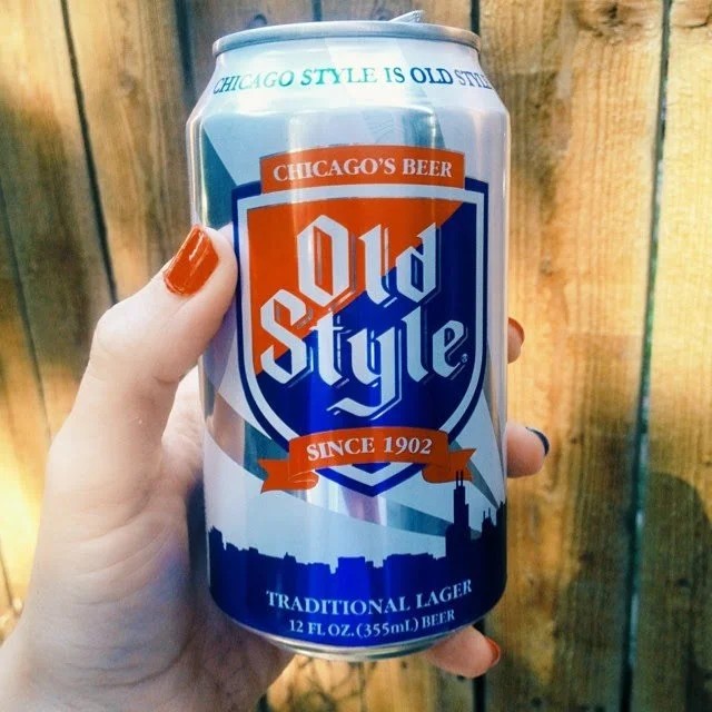 A person's hand holding a can of Old Style beer in front of a wooden fence.