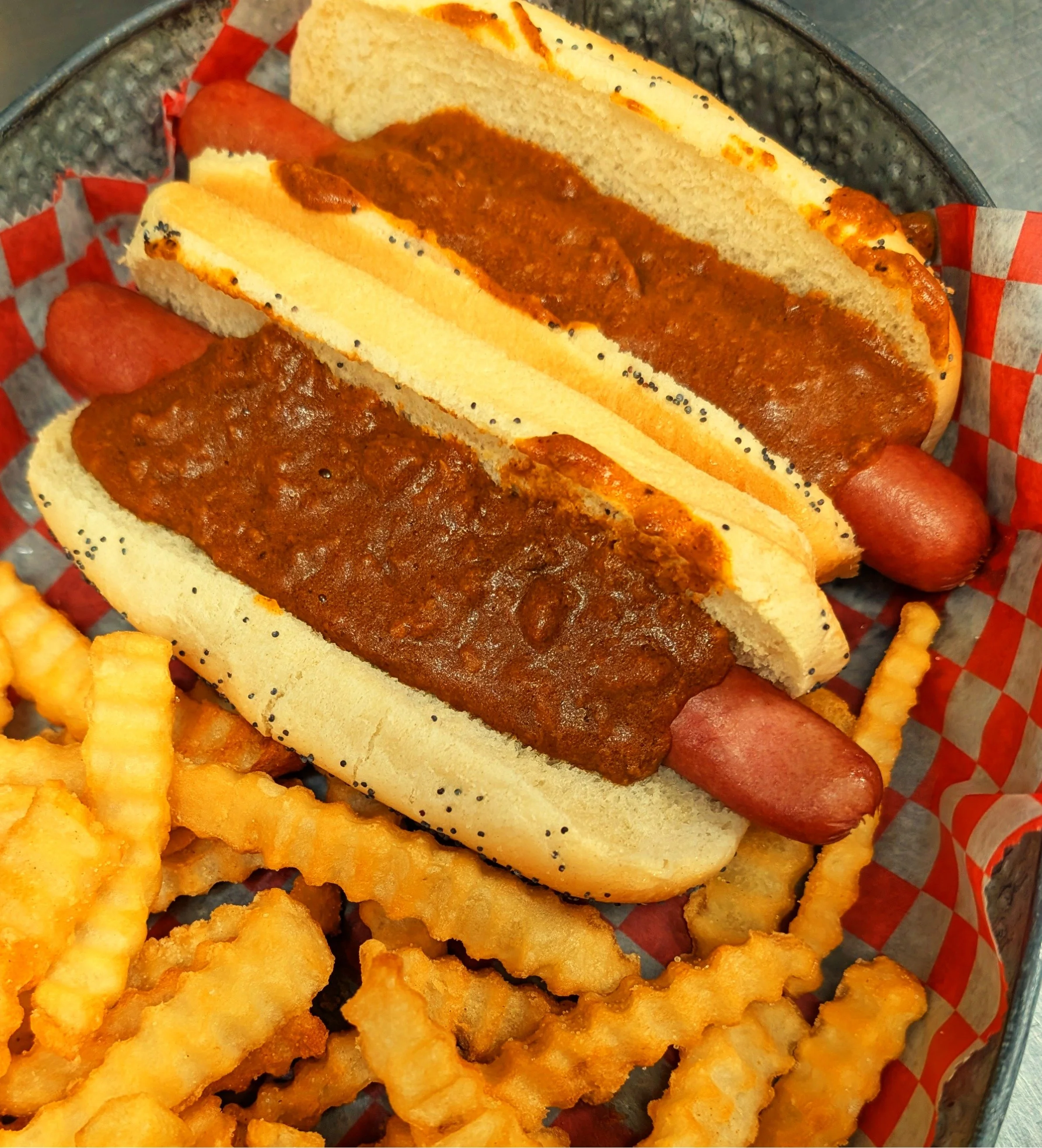 Hot dogs with grilled buns, chili, and mustard served with crinkle-cut French fries in a basket.