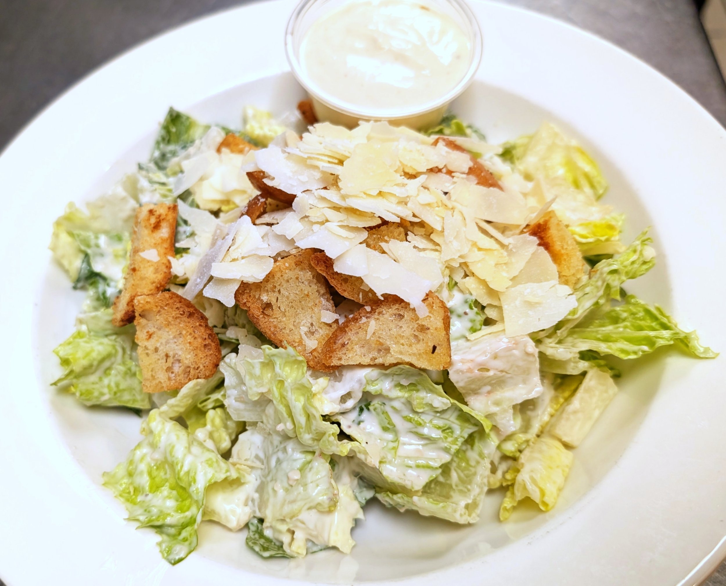Caesar salad with romaine lettuce, croutons, shaved Parmesan cheese, and a small cup of creamy dressing on a white plate.