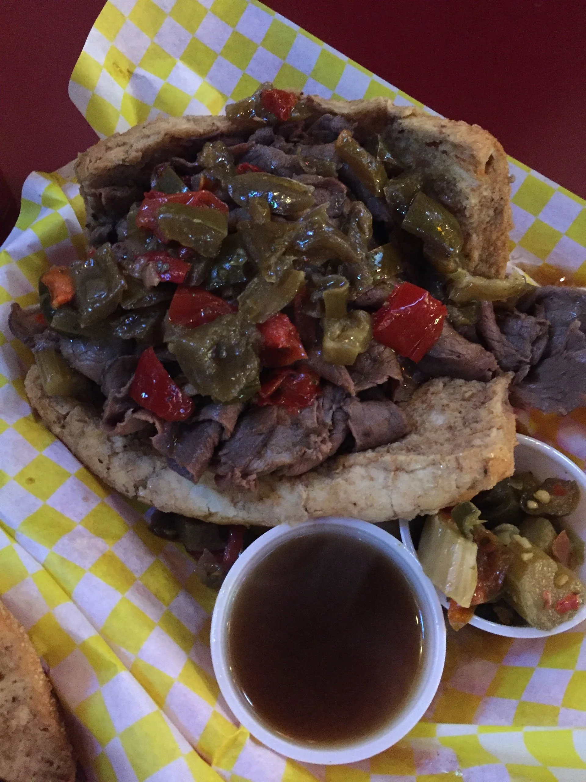 A sandwich with sliced beef topped with jalapeño peppers and red peppers, served in a bun, with a side of dipping sauce and pickled vegetables on yellow checkered paper.