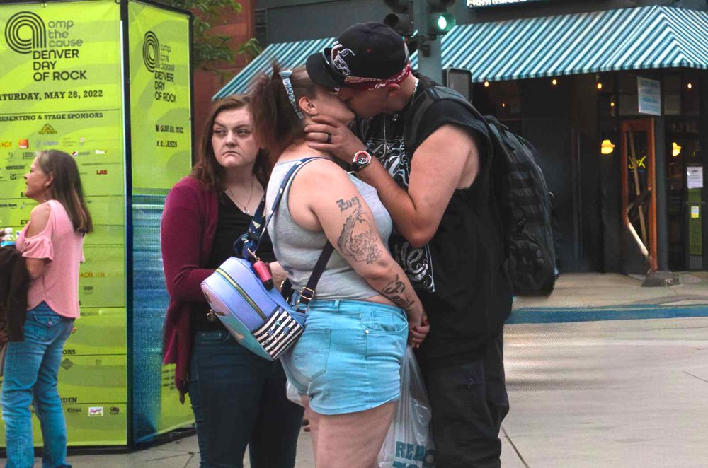  A guy, who was holding hands with both the woman in the grey tank top and the woman in the red sweater, kisses his girlfriend while the other woman looks away in displeasure. 