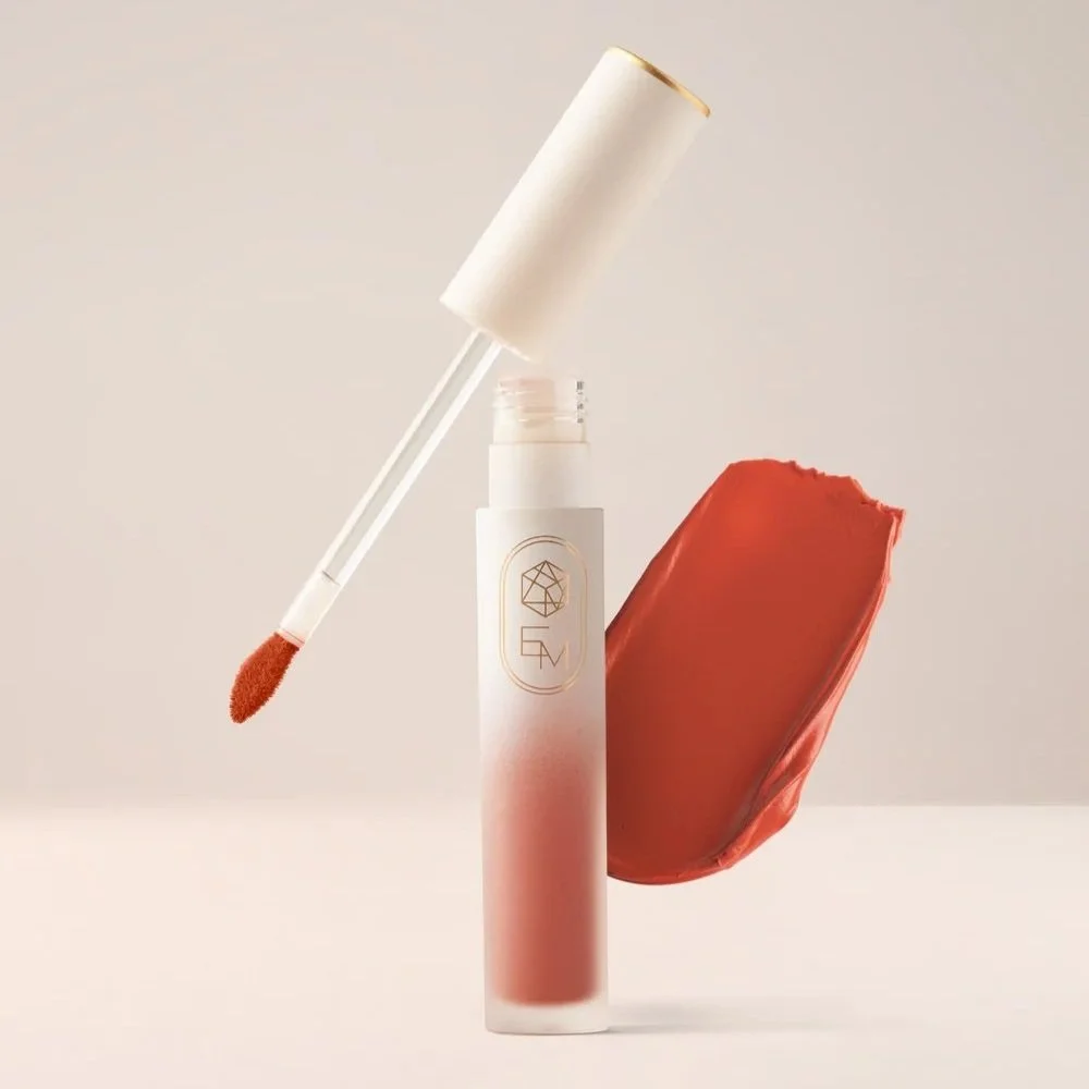 Cosmetic product with a white and pink gradient bottle, a lip gloss applicator with a bright reddish lip gloss, and a red object or bag in the background.
