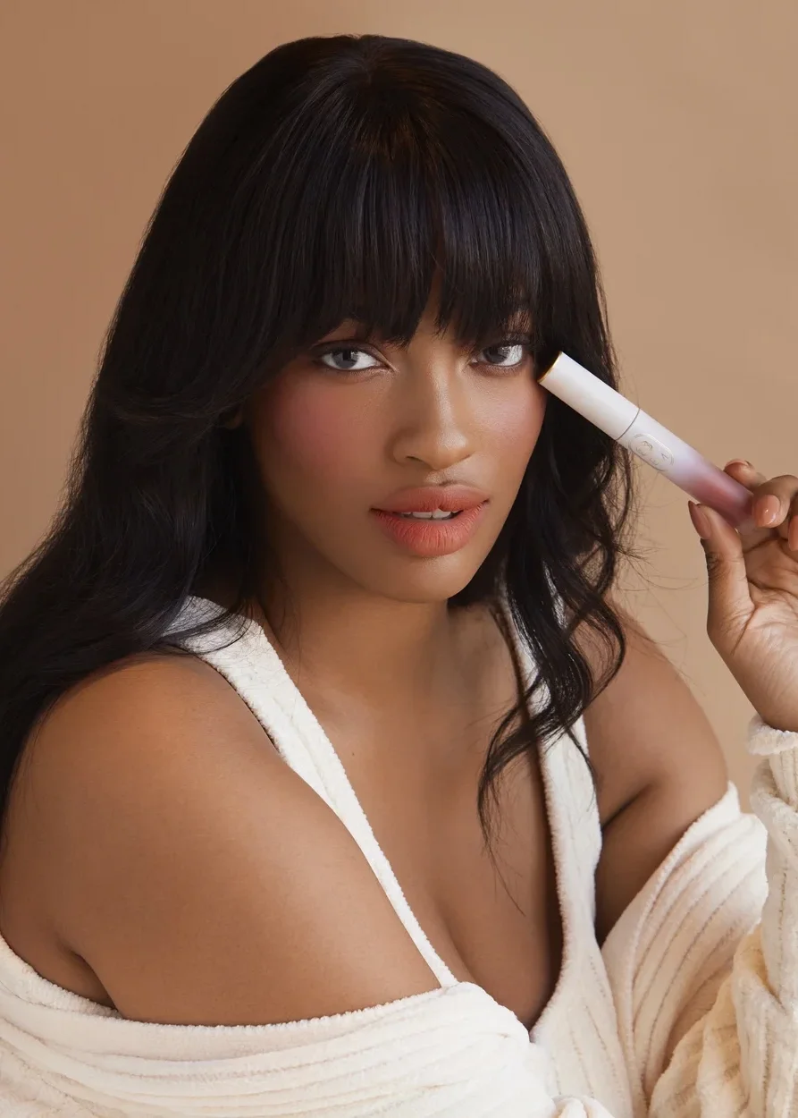 A woman with dark hair and bangs holding a skincare device near her face, wearing a white off-shoulder top, with a neutral background.