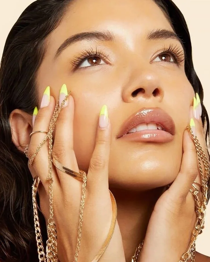 A woman with long dark hair, wearing gold jewelry and with yellow and white painted nails, touching her face and looking upwards.