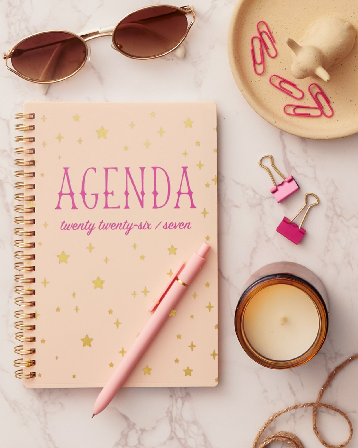 Pink and gold agenda with yellow stars and pink writing, pink pen, sunglasses, pink paper clips, pink binder clips, candle, and some jewelry on a marble surface.
