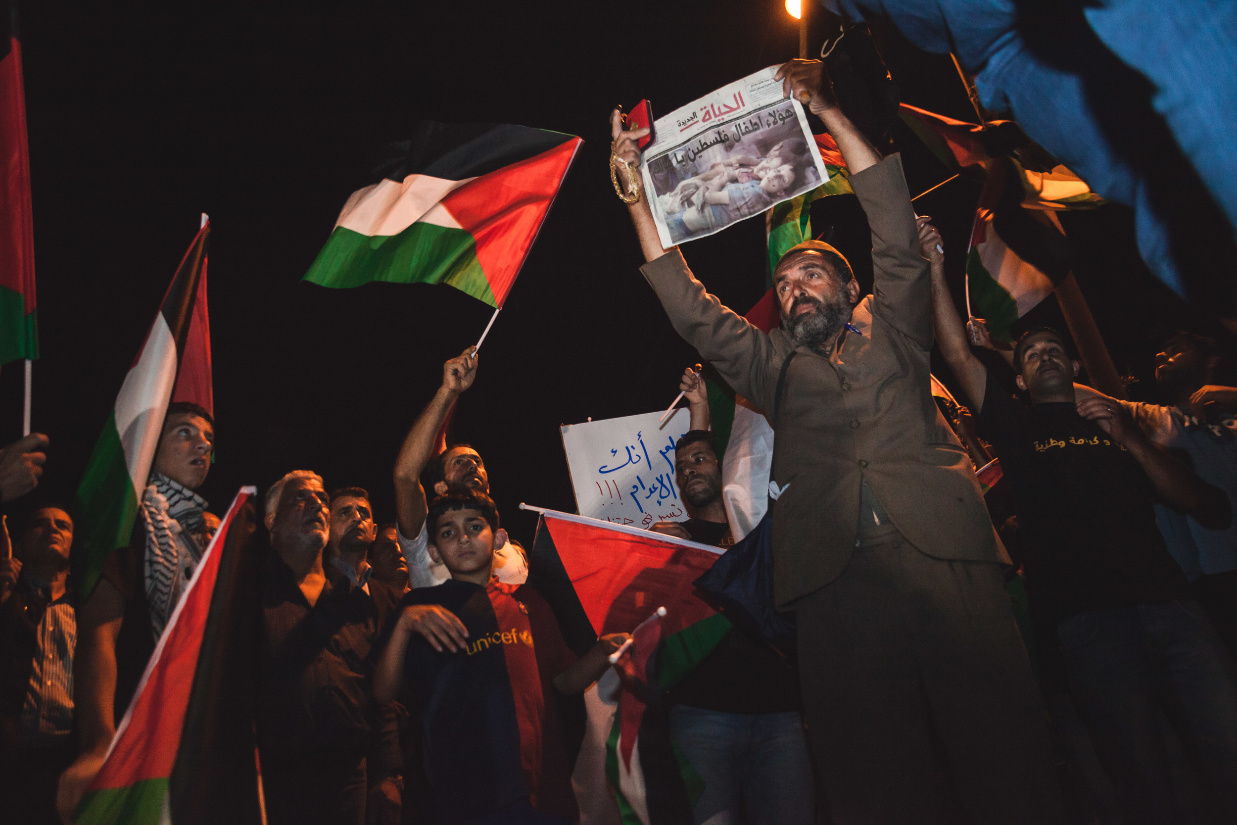  While the evenings protest was still in its early stages, a demonstrator holds up a newspaper with an image from Gaza prominently displayed as thousands gathers&nbsp;around the main junction of Al-Amari Refugee Camp waiting for the demonstrations to