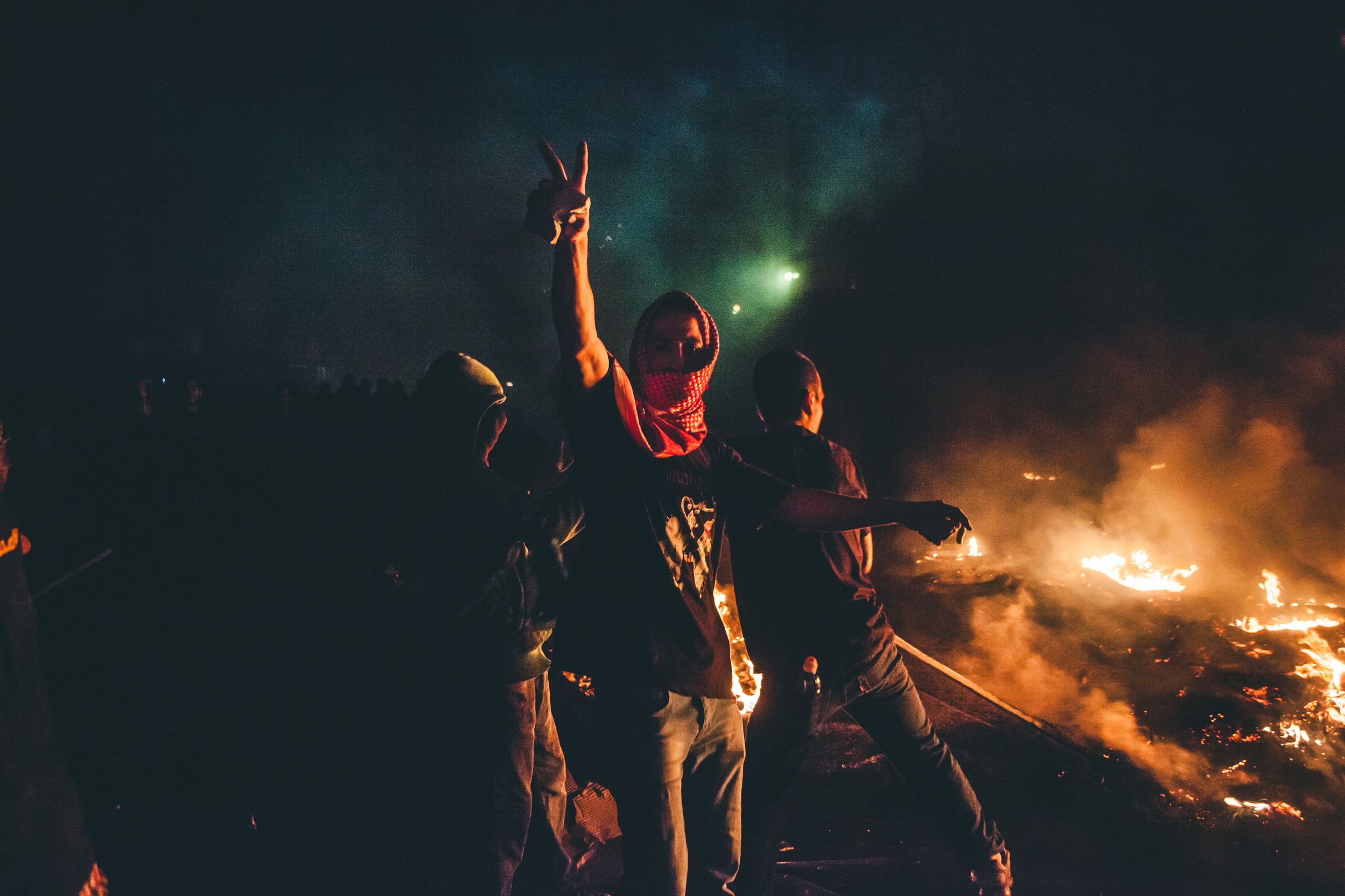  A keffiyeh clad demonstrator holds up a peace sign while clutching a rock&nbsp;at Qalandia checkpoint&nbsp;as the fire and protest roars behind him&nbsp;during the&nbsp;largest protest of the summer in the West Bank&nbsp;- the&nbsp;48Thousand March.