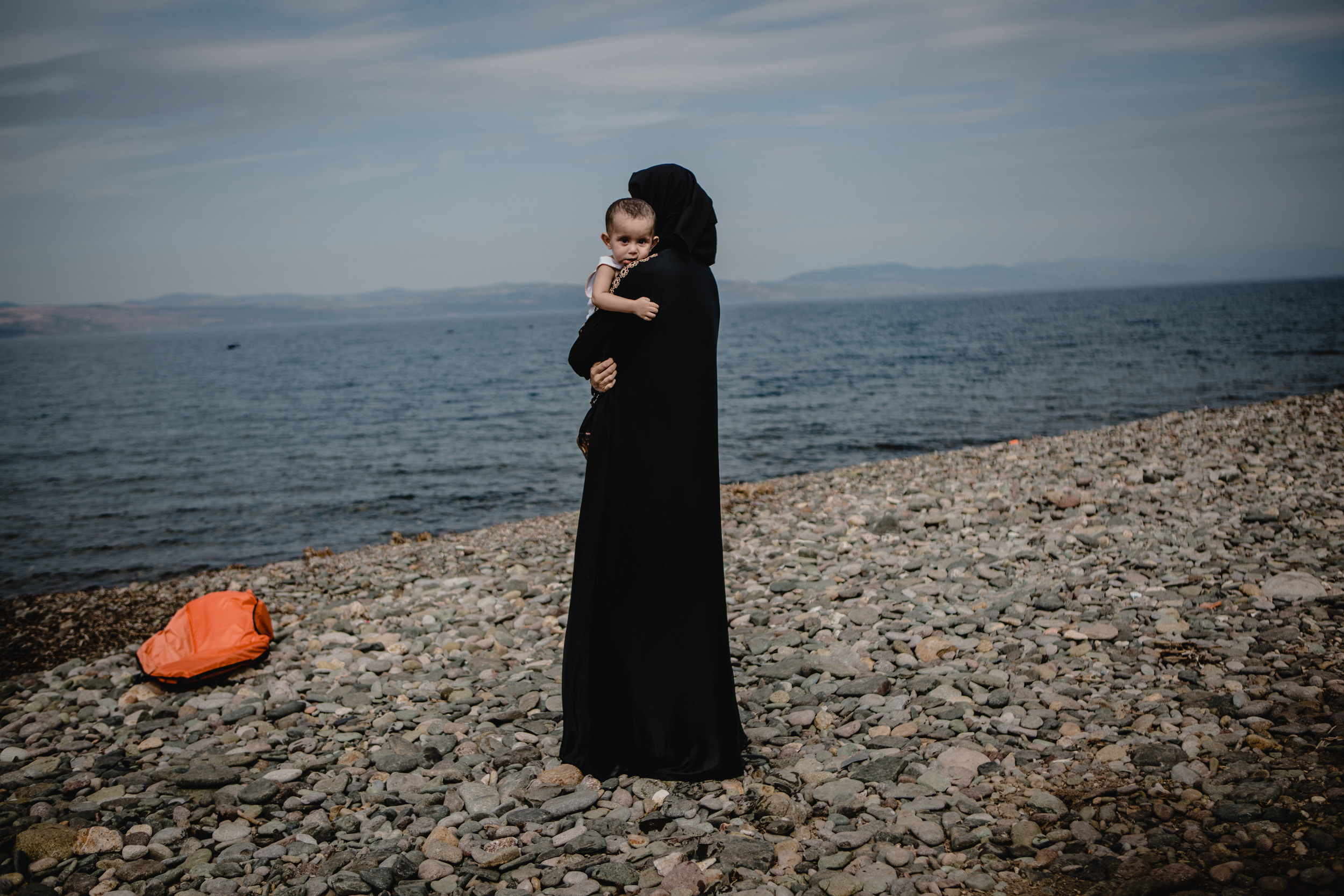 A women holds her child while looking back towards the sea she and her family just crossed from Turkey.