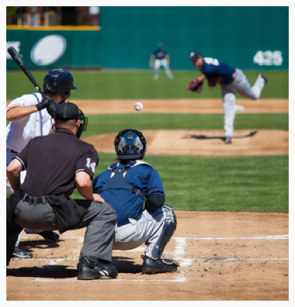 batter (left) in white uniform, umpire in black, catcher (crouching) in blue, pitcher (upper right) in blue.