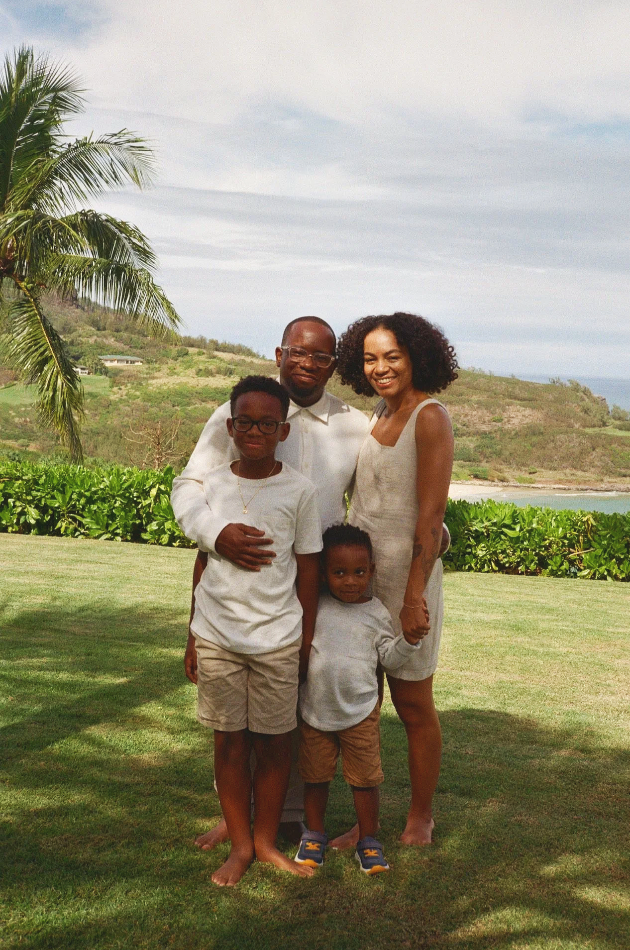 A family of four smiling outdoors with a tropical landscape in the background, including palm trees, green shrubbery, hills, and a body of water.