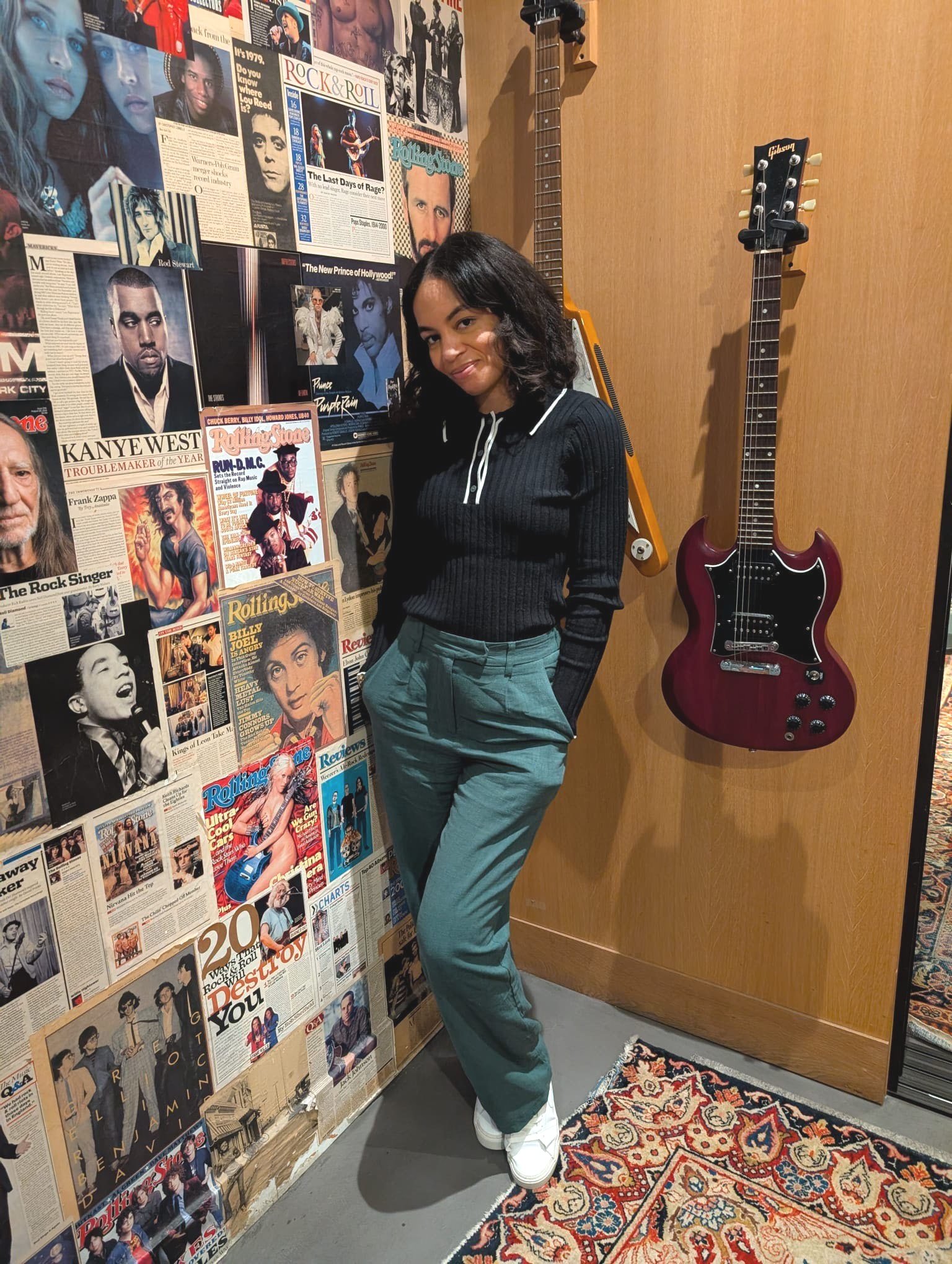 Woman standing indoors next to a wall decorated with music magazines and posters, with guitars mounted on the wall behind her.