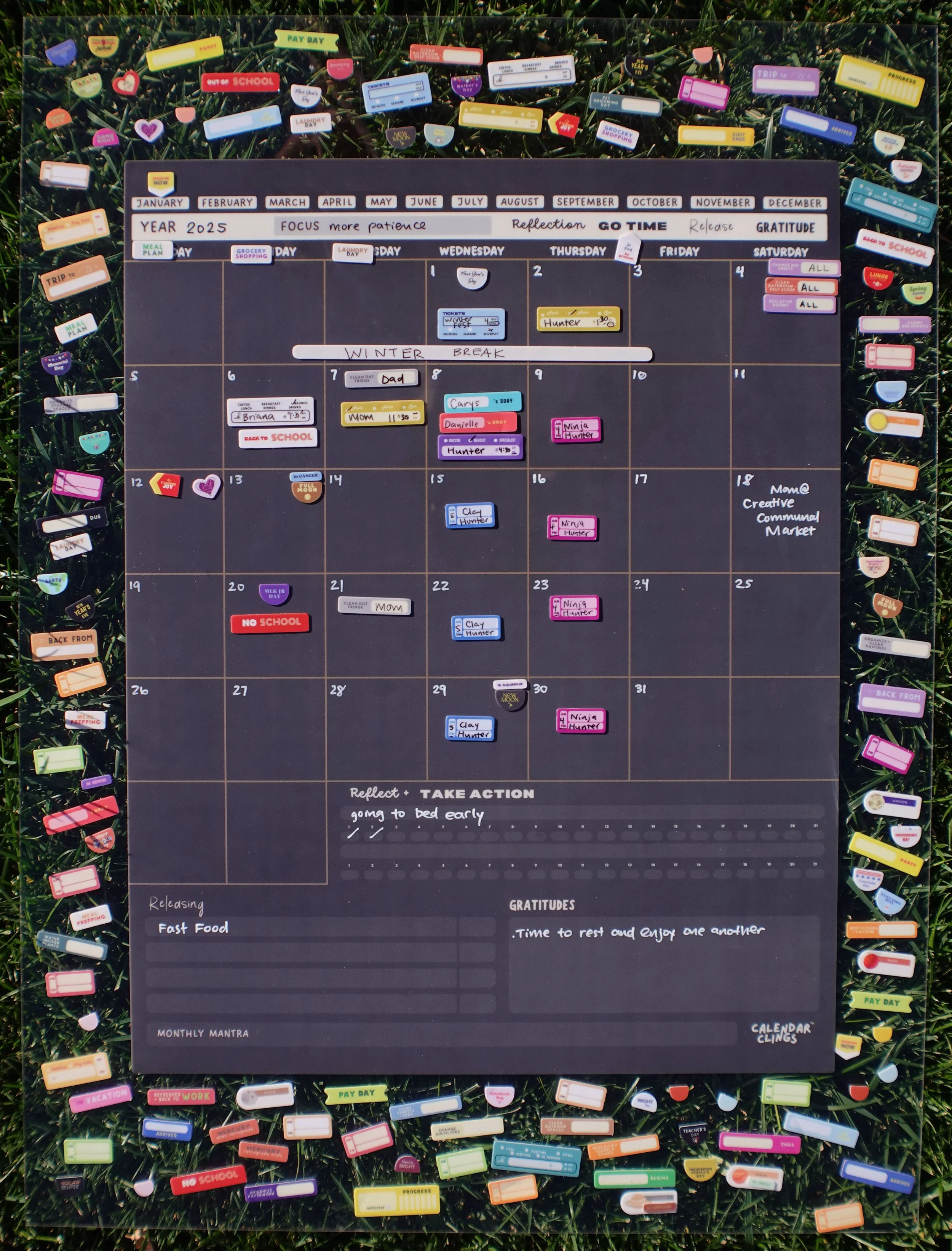 A calendar for January to December 2025, filled with personal appointments and events. It has motivational phrases at the top and reflection sections at the bottom. The calendar is decorated with numerous colorful labels and tags surrounding it, placed on a grassy background.