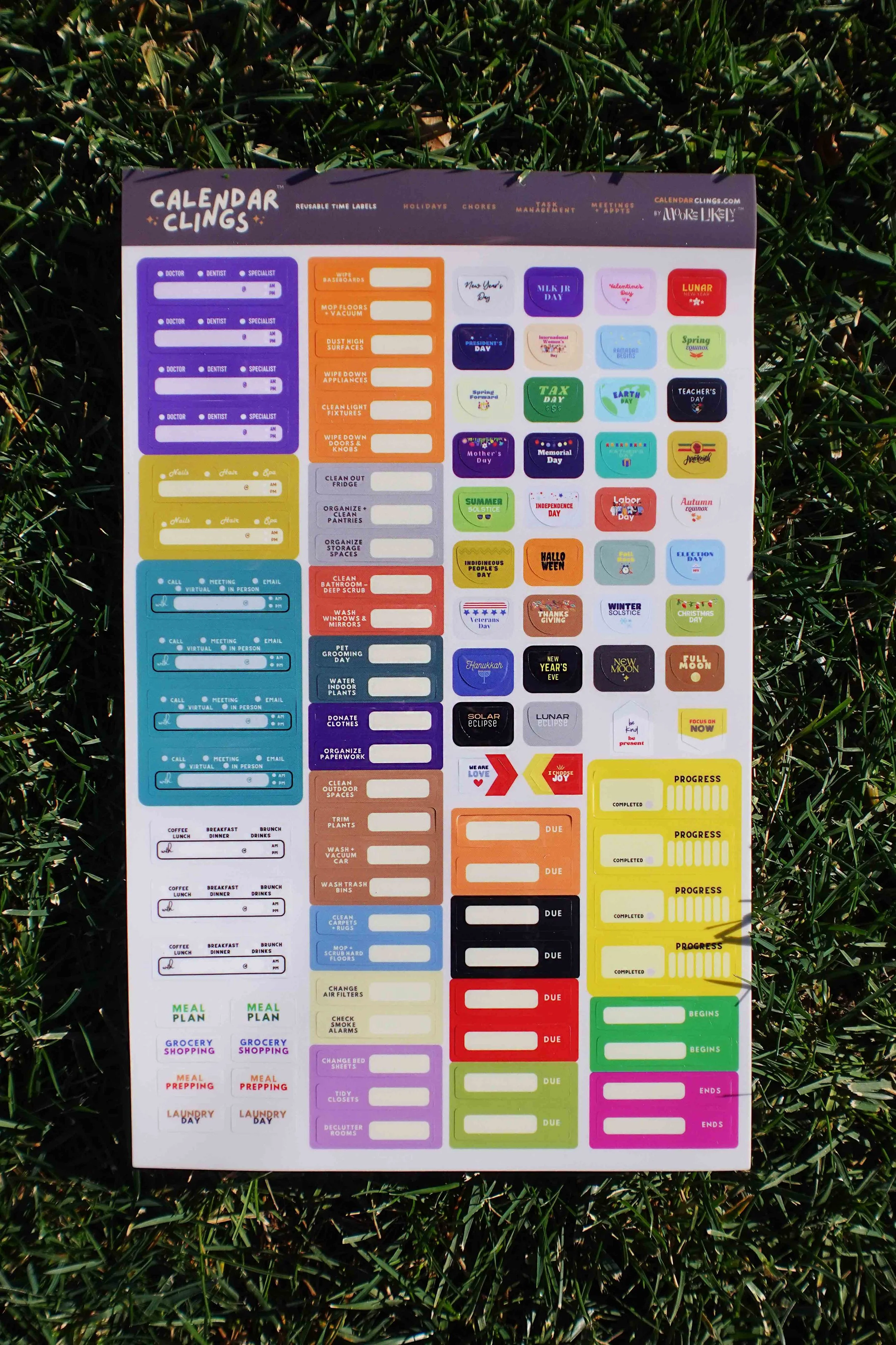 Colorful calendar cling stickers with labels for various dates, tasks, and holidays, including medical appointments, chores, work, holidays, and special events, on a sheet placed on grass.