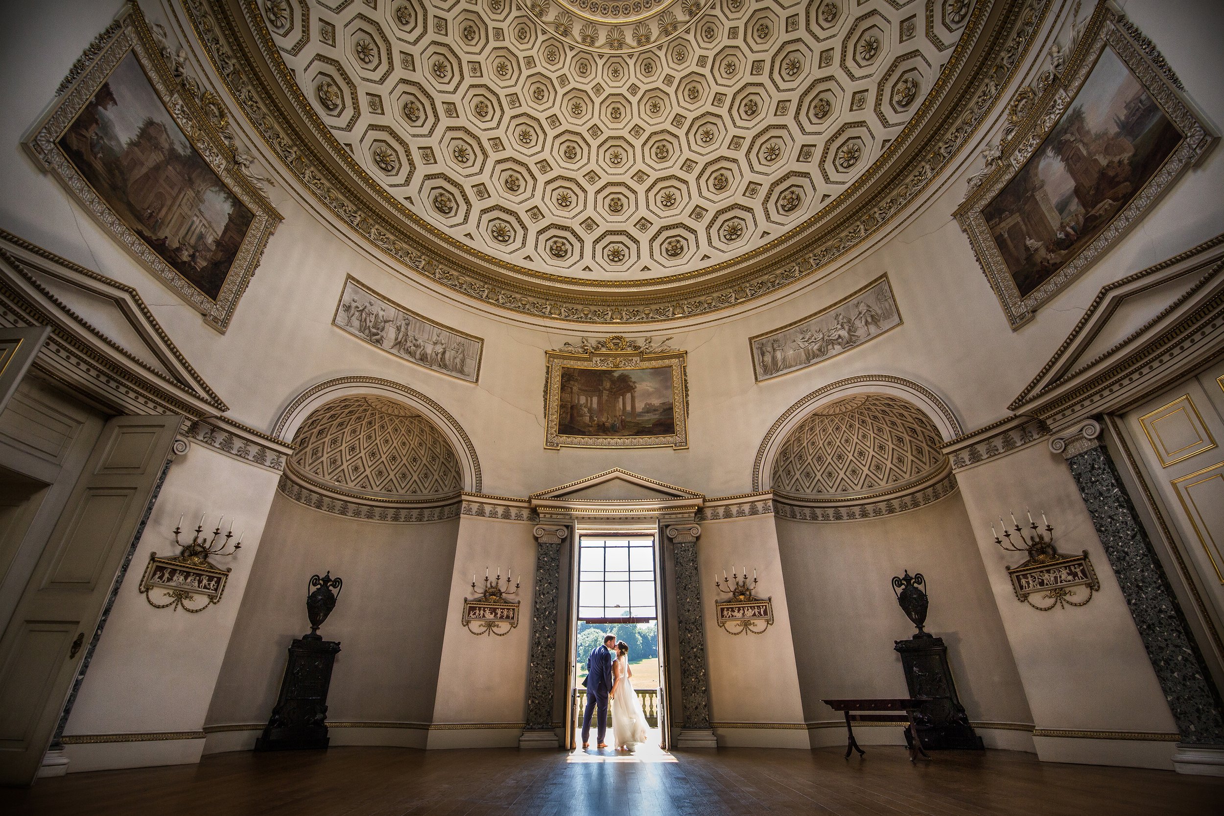 A bride and groom standing in a grand, ornately decorated hall with a domed ceiling, classical paintings and sculptures, and large windows.