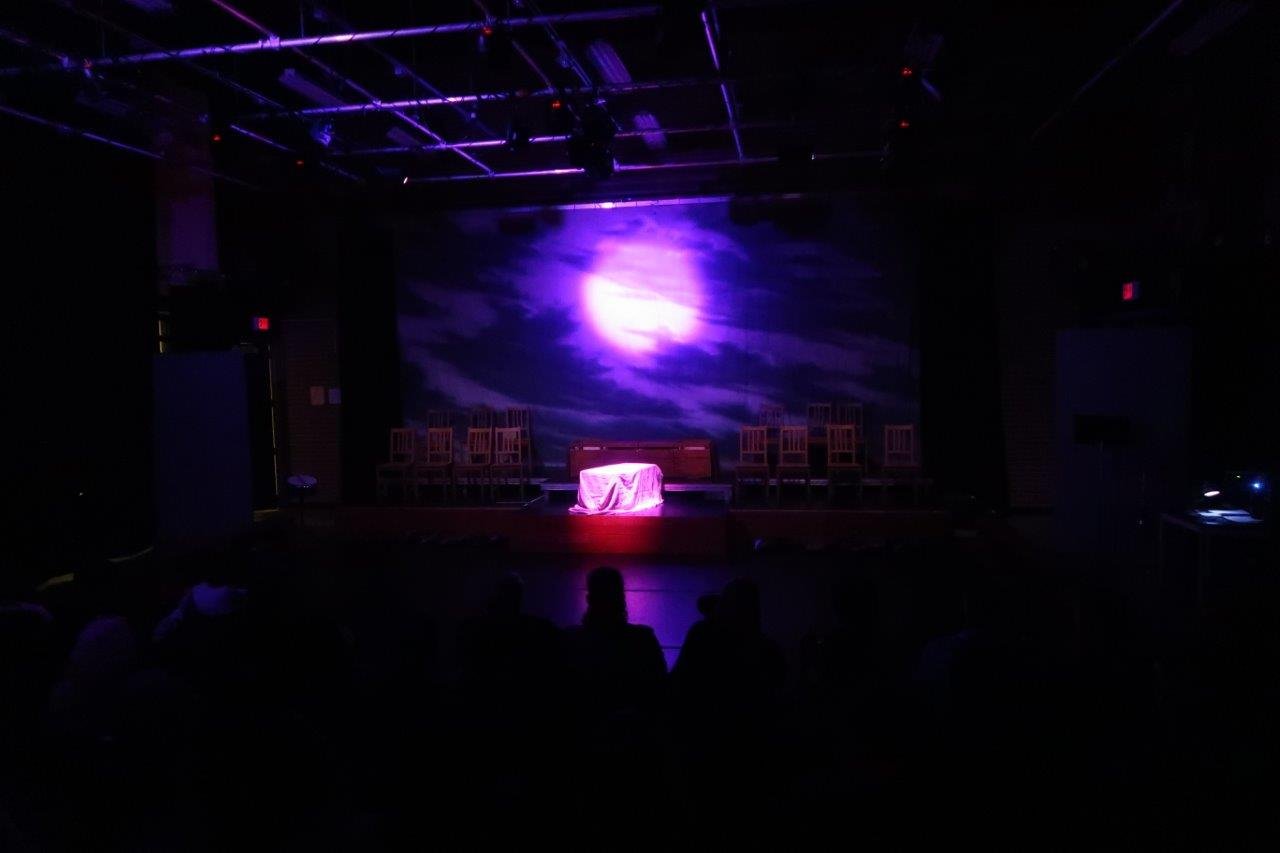  Stage lit with a full moon shrouded by clouds.  Chairs and a draped object in the middle. 