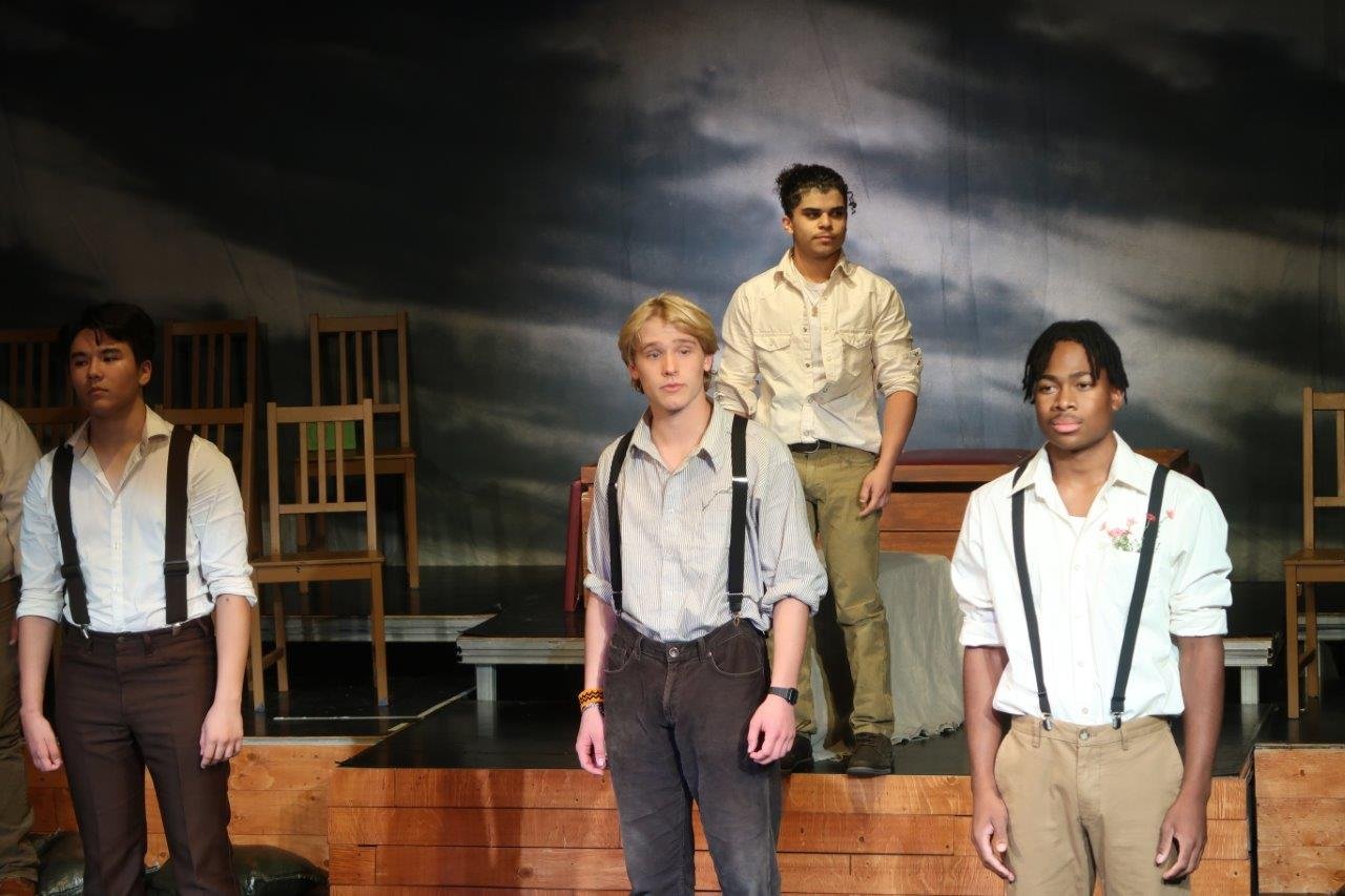  3 actors wearing white shirts and suspenders in the foreground, with 1 actor standing on a platform behind close to the backdrop. 