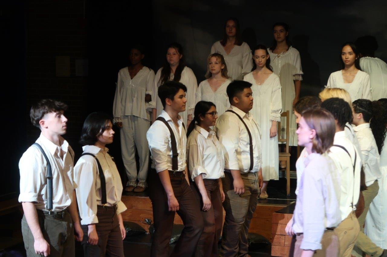  Two rows of actors facing each other in the foreground, as others look on from the raised platform in the back. 