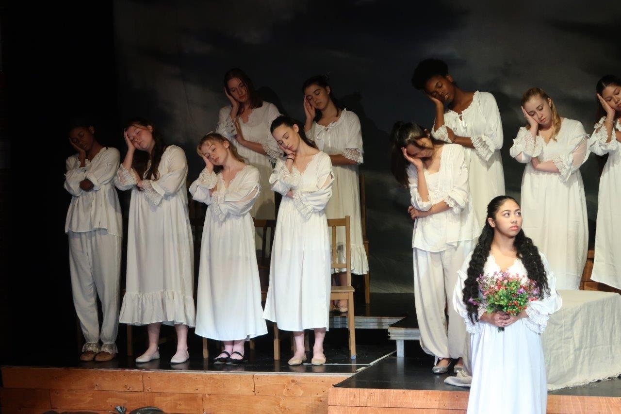  One actor dressed in white in the foreground holding a bridal bouquet, while the others on the back platform are standing while appearing asleep with their heads resting on their hands to their right. 