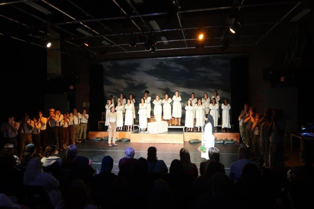  Scene of full stage, with actors in suspenders to the left and right, while others are dressed in white standing on a raised platform in the back.  Two actors are speaking to each other in the middle. 