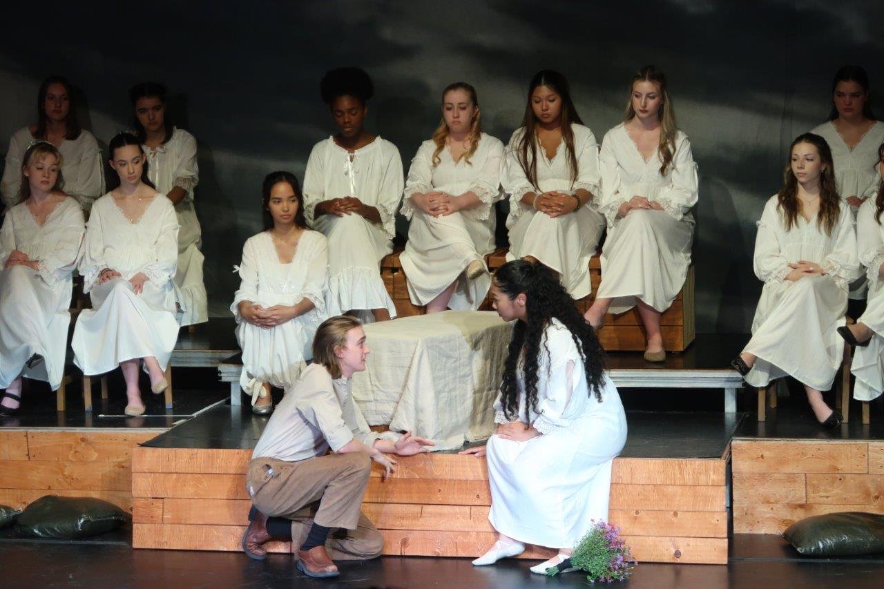  Two actors in the foreground crouched and looking to one another as they speak, while others on the raised platform watch them. 