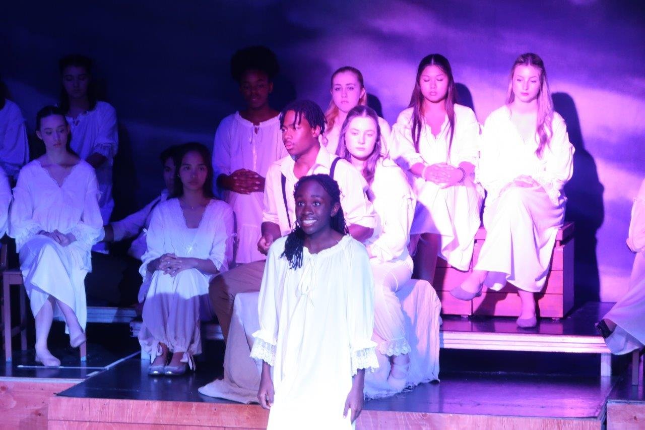  Actor in foreground spotlight speaking as others look upon them. 