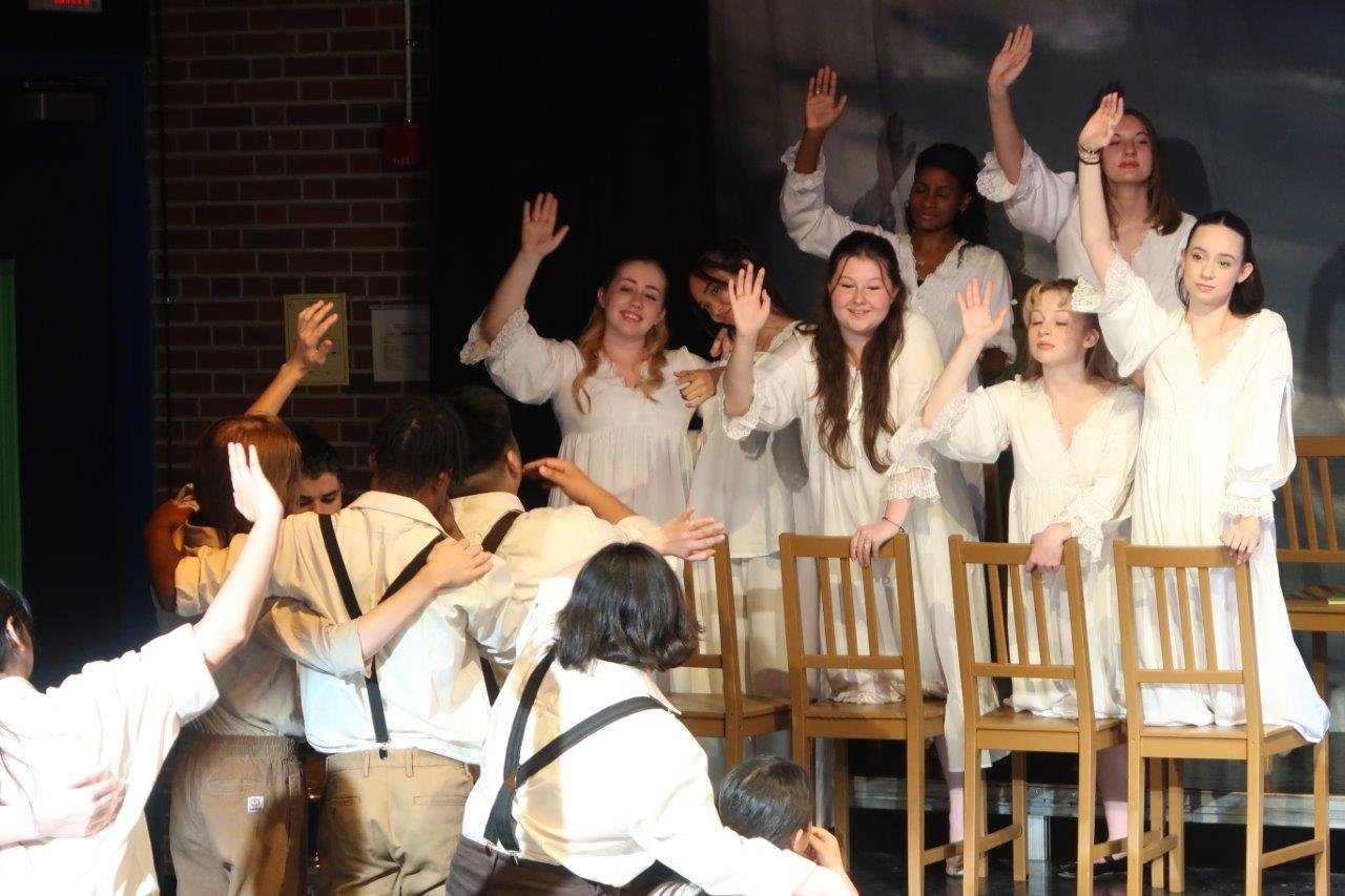  Foreground of actors in suspenders waving their hands with their backs to the picture towards others dressed in white waving back at them. 