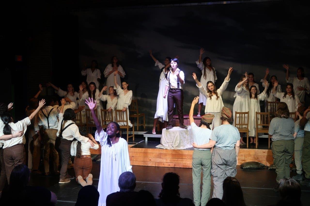  Foreground has an actor dressed in white with their right hand held high waving, with groups of actors waving to other groups. 