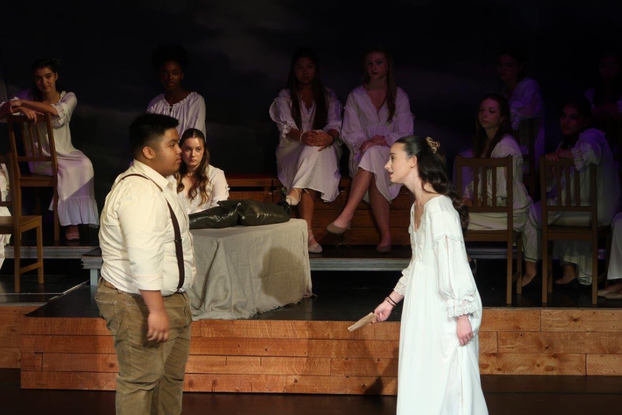  Two actors, one in white shirt and suspenders and the other in a white dress, speaking to one another in the foreground as others look on. 