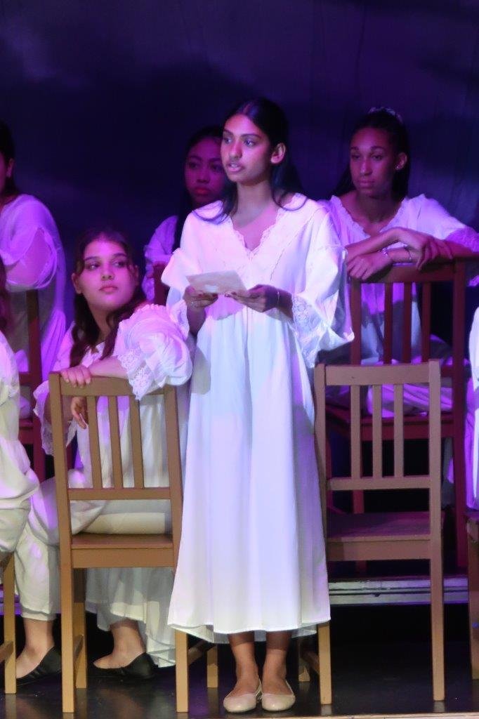  Actor in white dress standing and speaking as others look on seated behind them. 