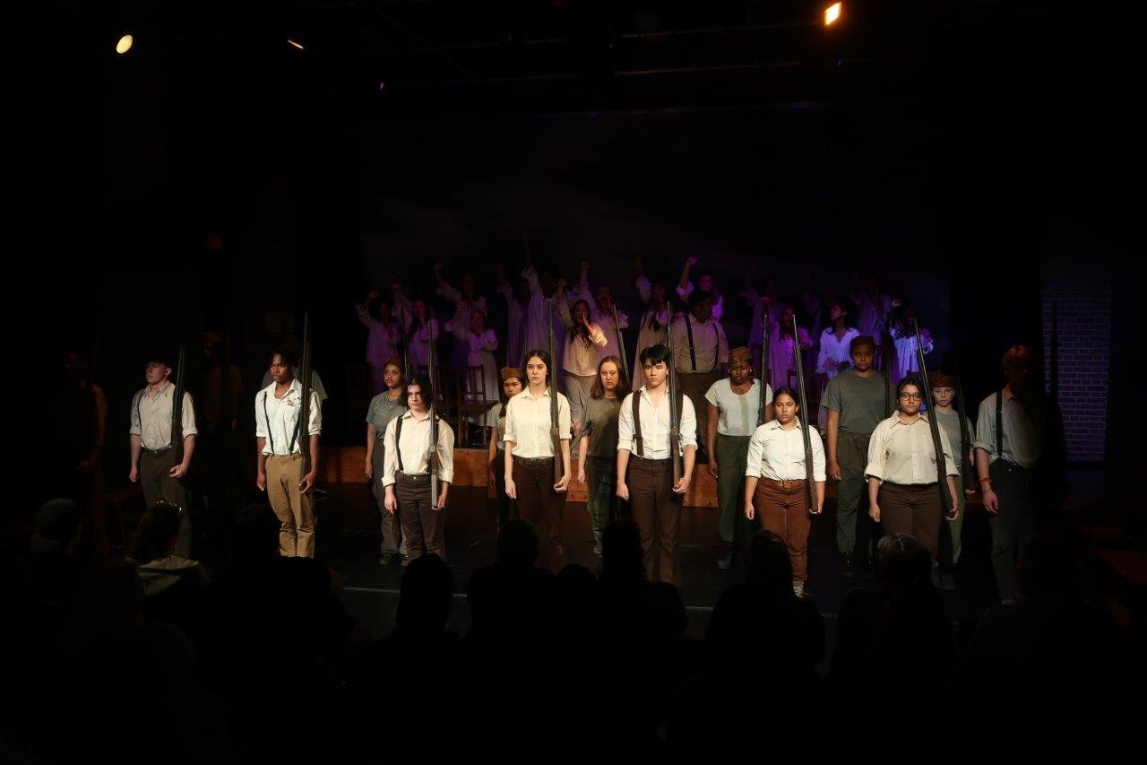  Row of actors in white shirts and suspenders in the foreground, with others standing in the background behind them. 