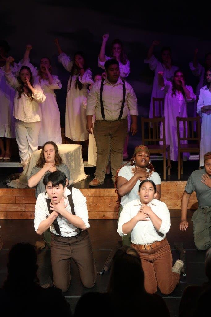  Actors in white shirts and suspenders in the foreground holding their hands to their necks as they lean forward, while others in the background look on. 