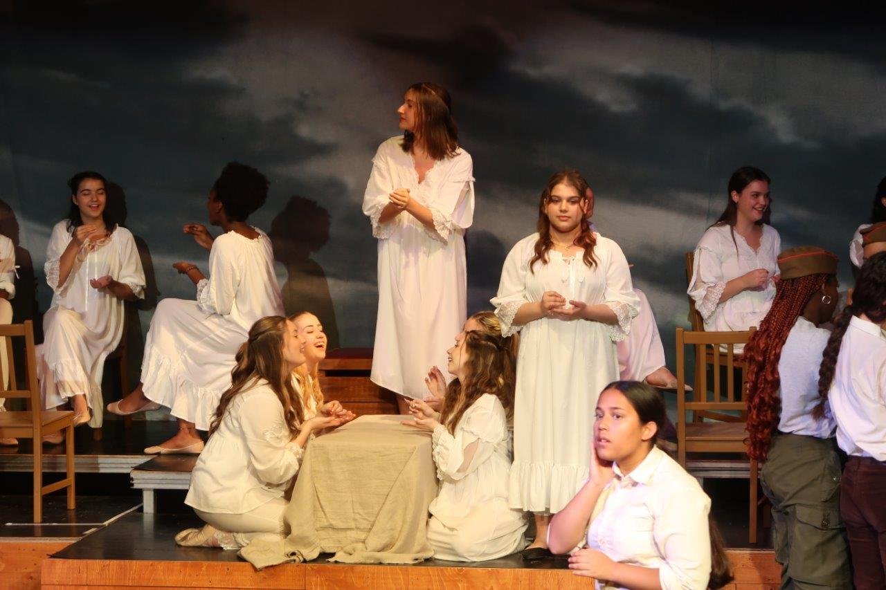  One actor in the front with a white shirt looking to their right as the background has a meal scene as others in dresses are eating or serving. 