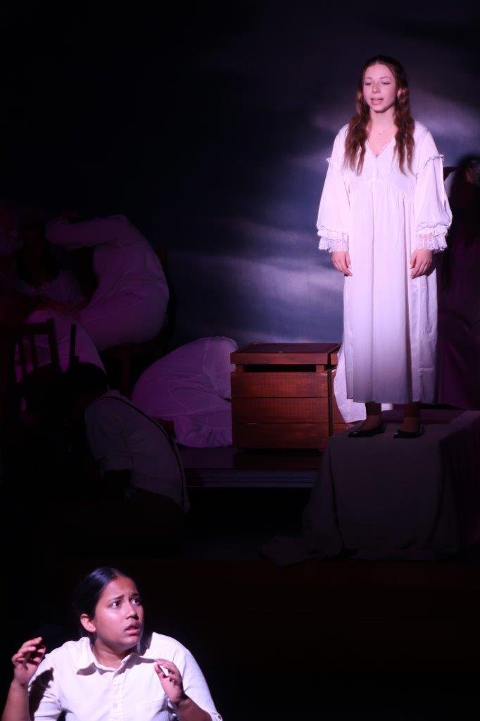  Foreground of actor in white looking shocked while another actor looks on them while speaking, spotlighted in their white dress. 