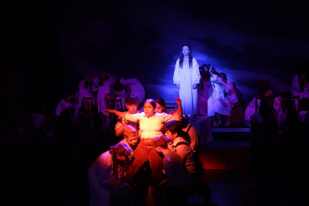  Orange spotlight on foreground of one actor in white shirt being lifted up by other actors, while in the background a blue spotlight of an actor looking upon them. 