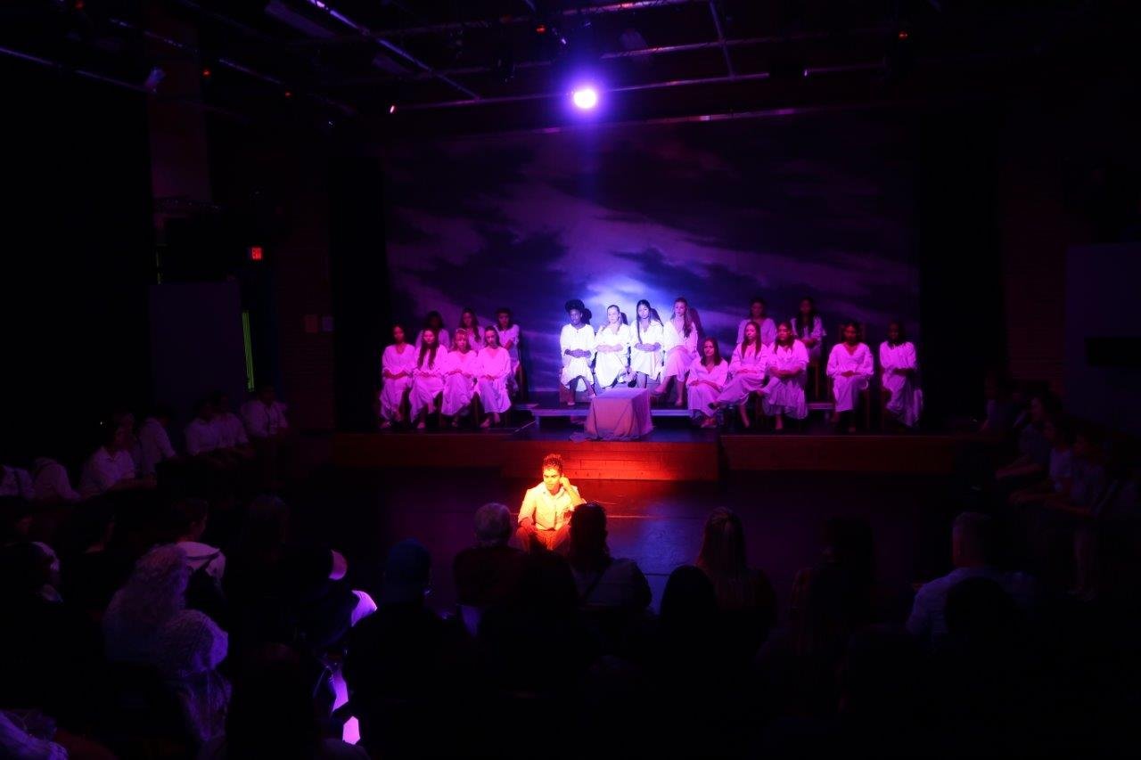  Whole stage scene of an actor sitting on the floor under an orange spotlight, while in the background a blue spotlight on a group of actors in the centre seated on a raised platform flanked by others seated on either side of them. 