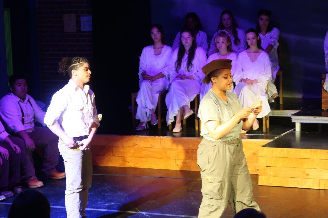  One actor in a soldier hat reading something out loud while another actor in white shirt looks at their back.  Others are looking upon them on a raised platform and seated behind them. 