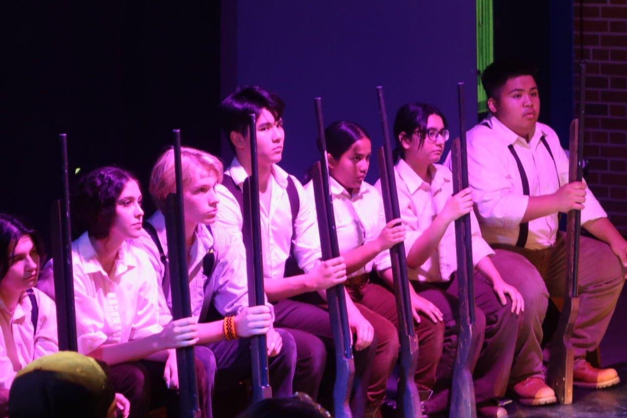  Row of seated actors wearing white shirts and carrying rifles 