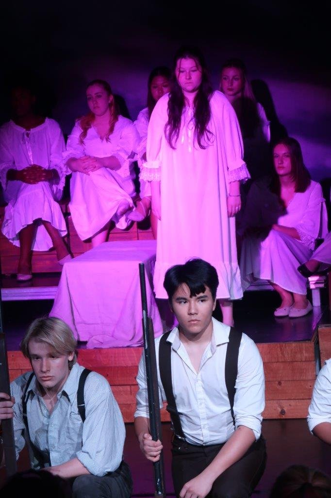  Two actors in white shirts and suspenders holding their rifles in the spotlight in the foreground, as a standing actor in a white dress looks on in the background. 