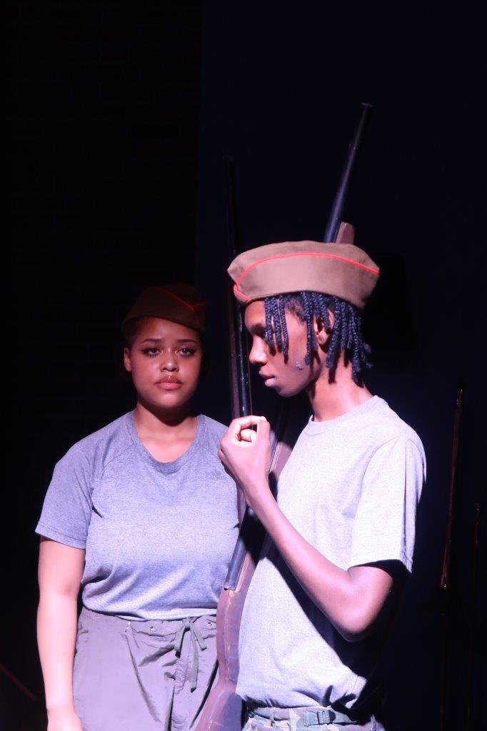  Actor with a soldier’s hat and carrying a rifle in their right hand raising his left hand to his lips looking to the left, as another actor carrying a rifle looks on them. 