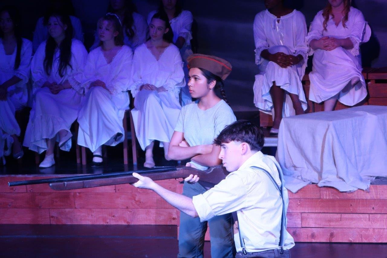  Actor wearing white shirt and suspenders crouched and aiming a rifle while a standing actor wearing a soldier’s cap follows their sightline.   